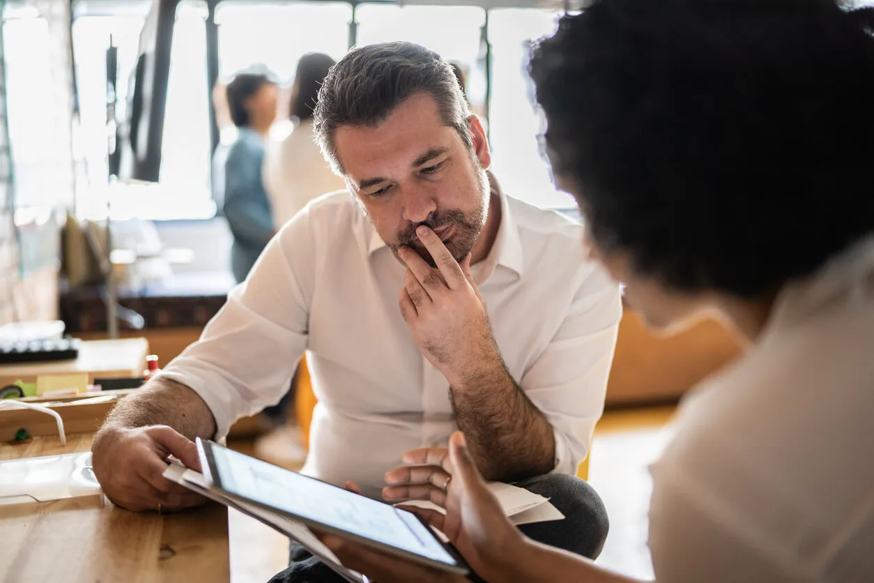 A man and woman sit at a table, engaged with a tablet, discussing something on the screen about b2b competitive analysis steps