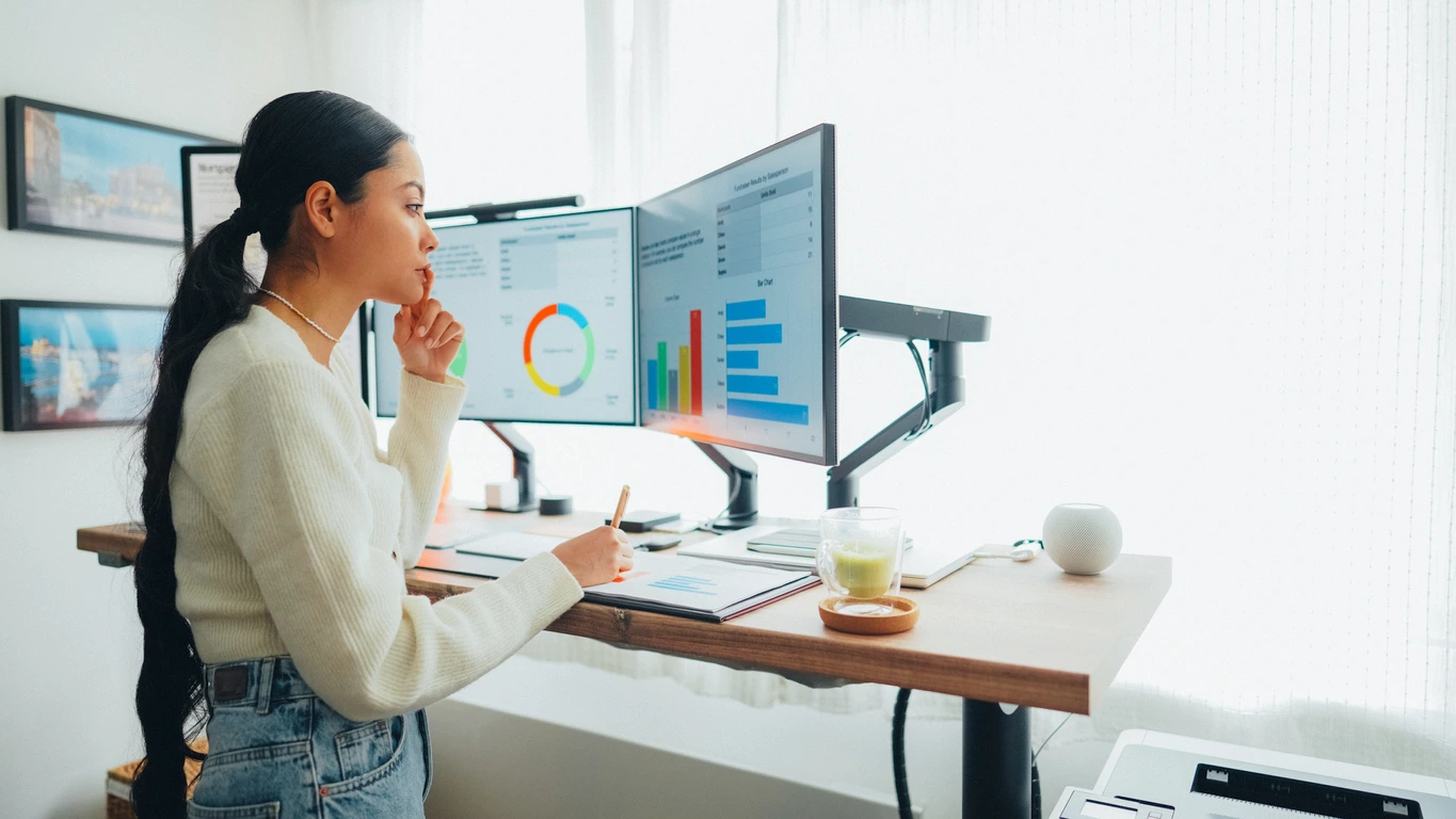 A woman sits at a desk, focused on two computer monitors displaying various applications and documents on how to improve b2b marketing performance