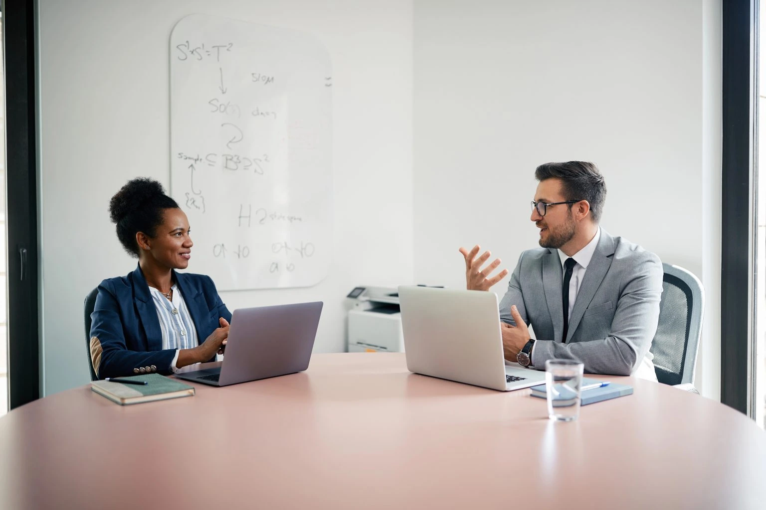 Two individuals seated at a table, each using a laptop, engaged in a collaborative work session discussing about the value of marketing in a b2b company