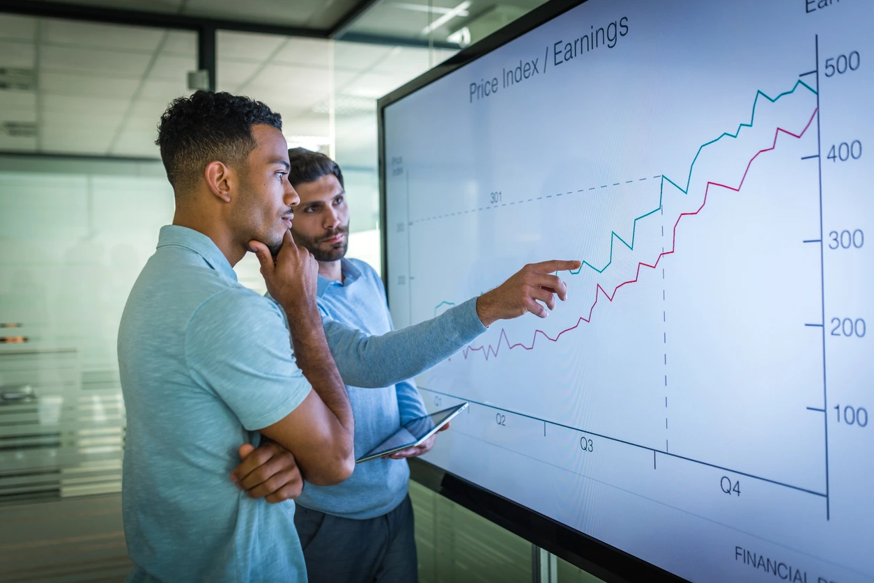 Two men engaged in a discussion, pointing at a chart displayed on a whiteboard while discussing b2b trends in marketing