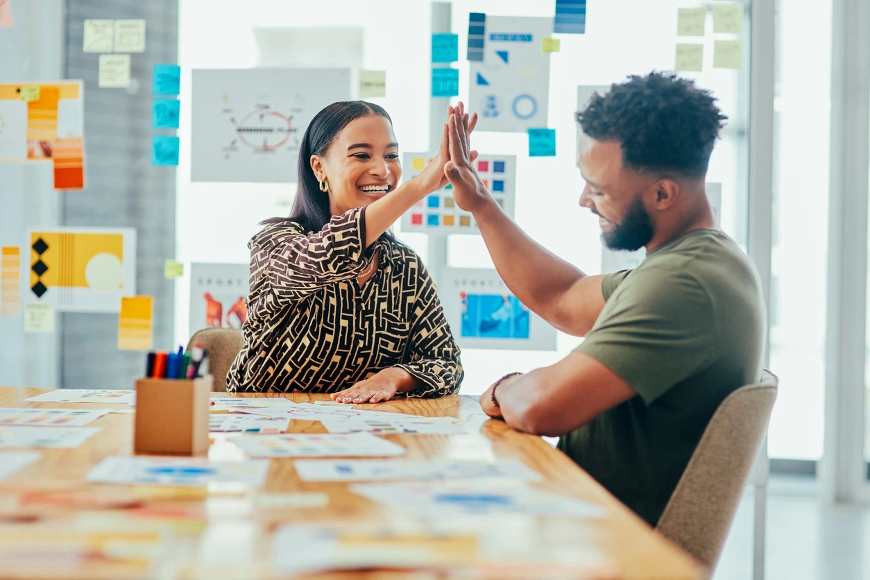 A man and woman enthusiastically high five each other while seated at a table, celebrating a shared achievements of b2b marketing competencies