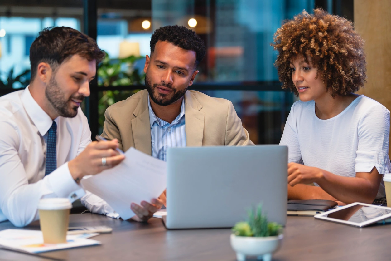 Three business professionals seated at a table, collaborating over a laptop during a meeting of b2b crm process